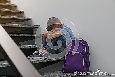 Upset little boy with backpack sitting on stairs indoors Stock Photo