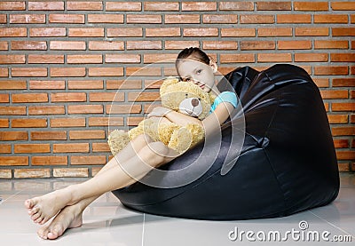 Upset caucasian teen girl sitting in black bean bag chair holding soft teddy bear toy against brick wall. Casual outfit. Child Stock Photo