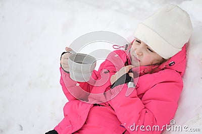 Upset frozen little cute girl in wool hat and pink ski suit drinking hot tea lying on white snow at cold winter holiday resort Stock Photo