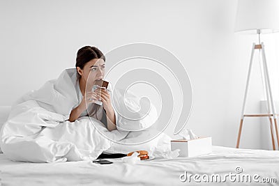 Upset hungry caucasian young woman sitting in bed at home and eating chocolate and junk food Stock Photo