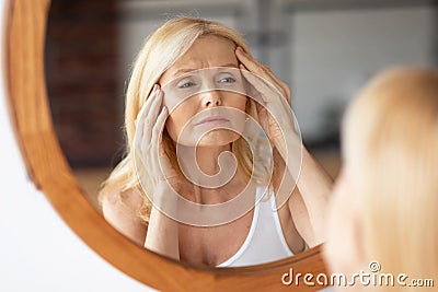 Upset middle aged woman looking at mirror and touching wrinkles on her face, lady examining fine lines on forehead Stock Photo