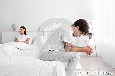 Upset offended caucasian young husband sitting in bed, suffering from weakness, wife ignoring boyfriend in white bedroom Stock Photo