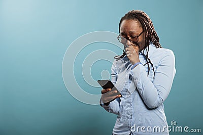 Upset young woman crying after reading tragic news on smartphone device while standing on blue background. Stock Photo