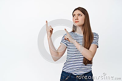 Upset young woman pointing and looking at upper left corner with skeptical, disappointed face expression, standing over Stock Photo