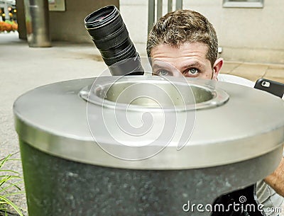 Lifestyle funny portrait of young paparazzi photographer man in action hidden behind city paper basket stalking for shooting excl Stock Photo