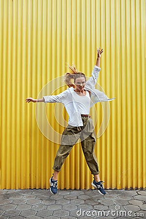 Urban. Fashion Girl Motion Portrait. Sexy Model In Casual Clothes And Glasses Against Yellow Iron Fence. Beautiful Woman Jumping Stock Photo