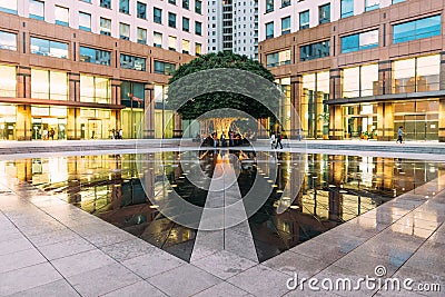 Urban space fountain area with big green tree in the corner with people. This area amid modern office buildings left and right. Stock Photo