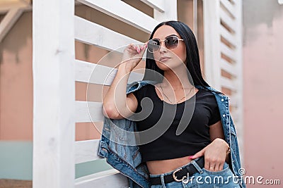 Urban stylish young woman hipster in a trendy black top in a fashionable denim jacket in a skirt in dark sunglasses posing Stock Photo