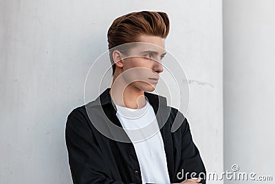 Urban young man with a stylish hairstyle in an elegant black shirt in a white T-shirt is standing near a vintage building Stock Photo