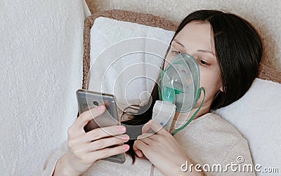 Use nebulizer and inhaler for the treatment. Young woman inhaling through inhaler mask lying on the couch and chatting Stock Photo