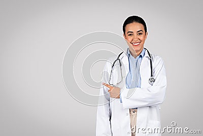 Useful information. Portrait of cheerful latin female doctor pointing at copy space, grey background, smiling at camera Stock Photo