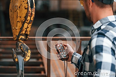 Using the controller with buttons to operate the little crane machine. Factory male worker in uniform is indoors Stock Photo