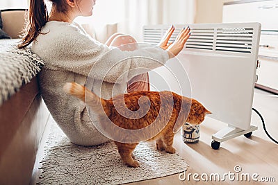 Using heater at home in winter. Woman warming her hands with cat. Heating season Stock Photo