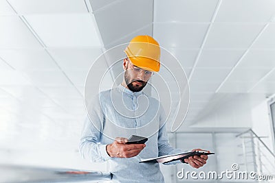 Using phone. Engineer in white clothes and orange protective hard hat standing and working indoors Stock Photo