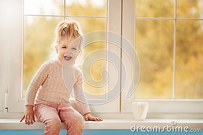 Ð¡ute little kid girl in pajama sitting by big window playing smiling enjoying home. Cacao cup standing on window. Autumn Season Stock Photo