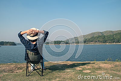 Asian woman wear hat and sitting relax on portable chair nearly lake at National Park. Stock Photo