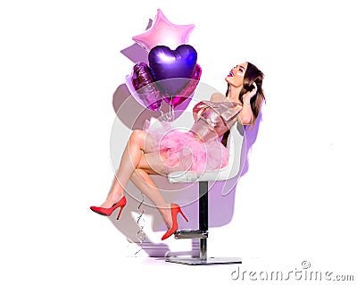 Valentine`s Day. Beauty fashion model party girl with heart shaped air balloons having fun, sitting on chair isolated on white Stock Photo