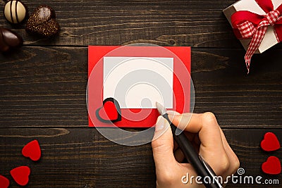 Valentine`s Day desk. Female hand with pen and Valentine`s Day card on the wooden background. Stock Photo