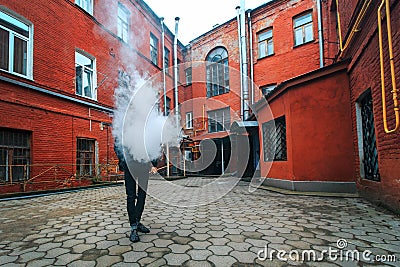 Vape man. A handsome young white guy in glasses blows steam from an electronic cigarette in a vintage old red yard. Stock Photo