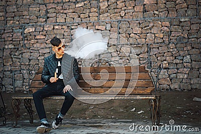 Vape. A young handsome guy sits on the bench and blows steam from an electronic cigarette. Stock Photo