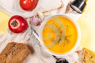 Vegetable cream soup in bowl, tomatoes, bread, garlic, spoon and spices on beige napkin on yellow backdrop. Top view Stock Photo