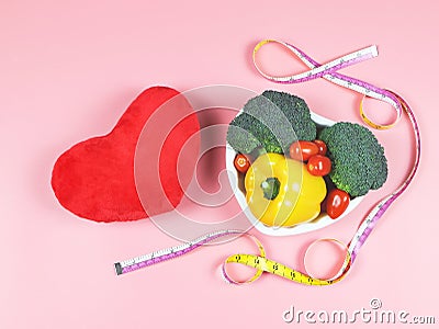 Vegetables capsicum, broccoli and tomato in heart shape plate, measuring tape and red heart shape pillow on pink background. Stock Photo