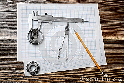 A vernier caliper holding a bearing, a pencil and a pair of compasses lying over drafting paper on wood background. Stock Photo