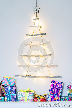 Vertical background of Unusual Christmas tree made of wooden sticks with festive lights on the white wall and colourful gift boxes Stock Photo