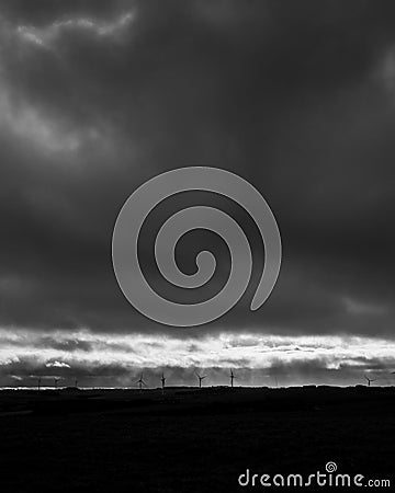 Vertical grayscale shot of dark thunder sky over silhouettes of wind turbines Stock Photo