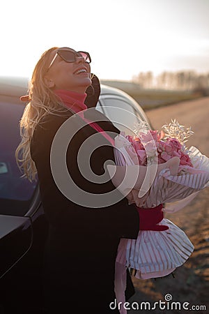 Vertical laughing fashionable blond woman in sunglasses holding posh pink rose flower decorative bouquet near black car Stock Photo