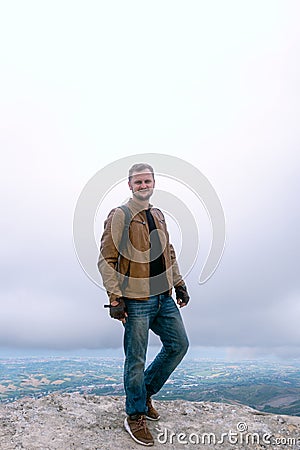 Vertical photo Portrait of a man dressed in a stylish brown leather jacket and jeans. backpack watches and biker gloves. posing on Stock Photo
