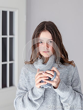 Vertical portrait of beautiful tween girl in warm grey pullover holding cup of tea looking at camera at home. Stock Photo