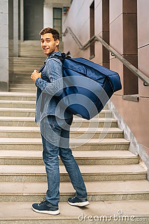 Vertical portrait of cheerful handsome young male of food delivery service with large thermo backpack standing posing in Stock Photo