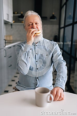 Vertical portrait of hungry mature elderly male eating fast food enjoying dinner at home and drink coffee. Hungry senior Stock Photo