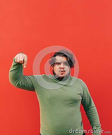 Vertical shot of an attractive emotional hispanic man standing on a red background, pointing his finger down, frowning, feeling Stock Photo