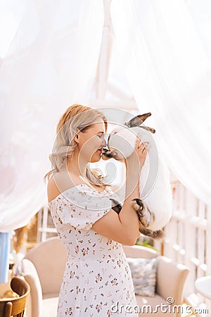 Vertical shot of happy blonde young woman in summer dress holding hugging cute fluffy white bunny standing inside of Stock Photo