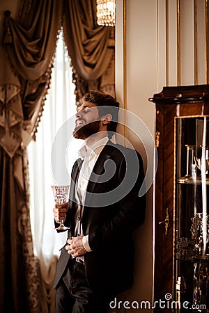 Vertical shot of pleased bearded young businessman wears black formal suit holds glass and drinks beverage, stands in Stock Photo