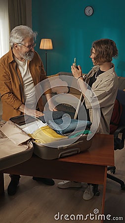 Vertical shot of a teenager, young man packing his belongings in a suitcase. His grandfather, granddad is helping him Stock Photo