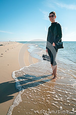 Vertical shot of a woman in dark outdoor clothes with shoes in her right hand, standing barefoot on a beach, with tide Stock Photo