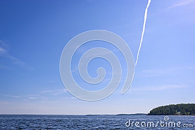 Vertical trail from the plane against the blue sky with cirrus clouds above the water surface of the lake Stock Photo