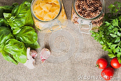 Various types of pasta, fusilli and farfalle, in glass jars next to tomato and garlic and basil Stock Photo