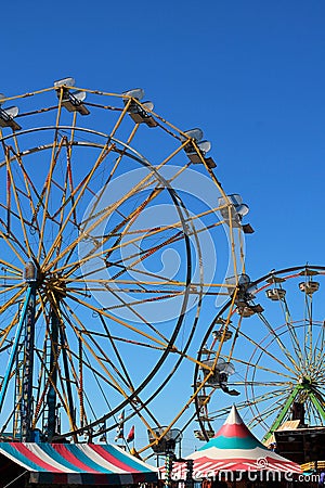 Vertical view of two colorful Ferris wheels in silhouette against a bright blue summer sky with tent pavilions below Editorial Stock Photo