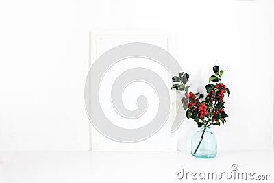 Vertical white blank wooden frame mockup. Holly berry branches in blue glass vase on white table. Styled stock feminine Stock Photo