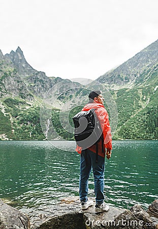 Vertical. Young tourist standing with his back on the shore of a beautiful lake in the rain on a background of mountains, wearing Stock Photo