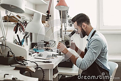 Very close. Side view of a male jeweler looking at the new jewelery product through microscope in a workshop. Stock Photo