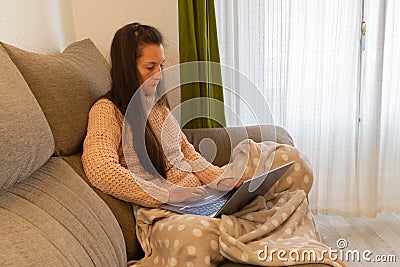 Very concentrated and serious woman sitting on a sofa and covered with a blanket works on her laptop from home Stock Photo