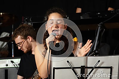 Close up face beautiful young brunette, vocalist singer with microphone, while singing live, with young guitarist in background Stock Photo