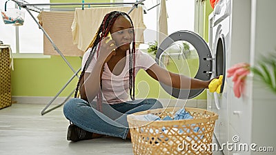 Vibrant african american woman passionately engaged in conversation on smartphone while washing clothes in busy home laundry room Stock Photo