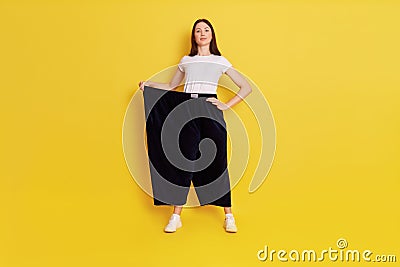 Victorious woman holding her too big pants and looking directly at camera, being proud of her result, standing isolated over Stock Photo