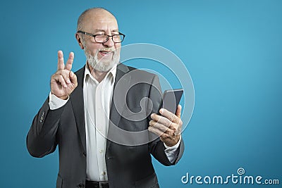 Video call by phone. Handsome elderly man greets a friend using a mobile phone in a classic suit on a blue background. studio Stock Photo